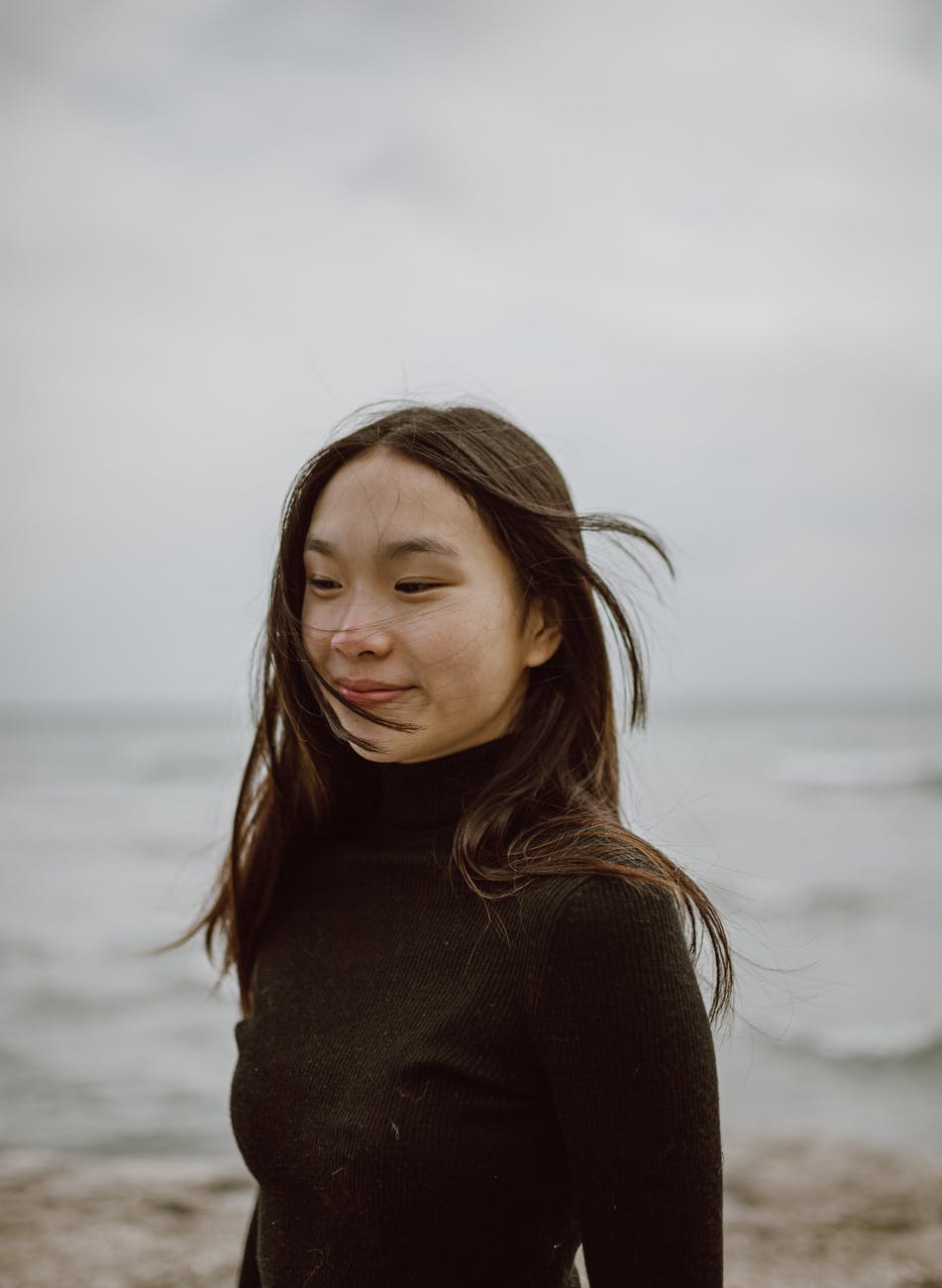 smiling young ethnic female traveler enjoying cold windy day on ocean beach