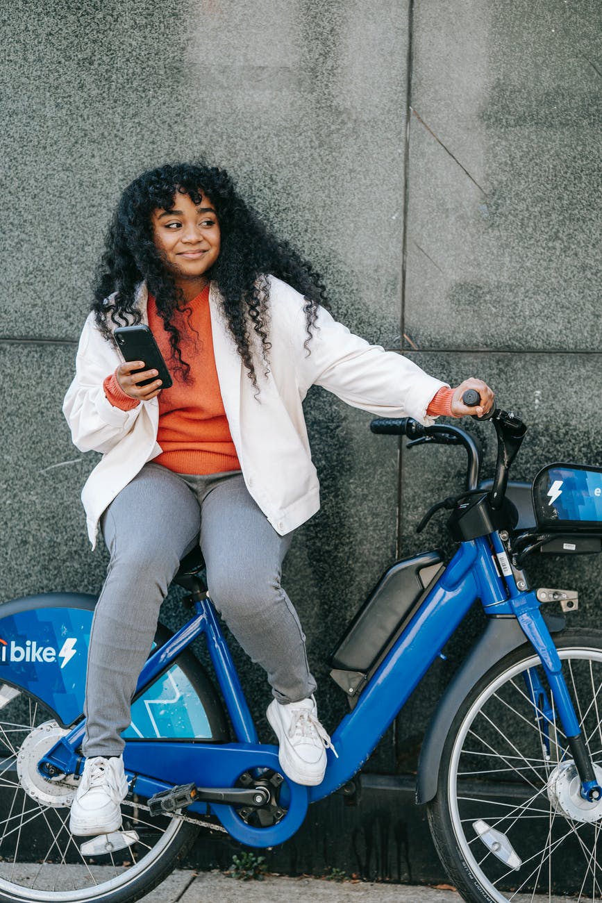 cheerful black woman with smartphone sitting on bicycle
